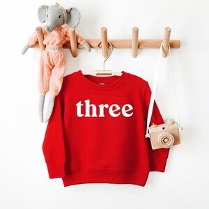 Puede incluir: Sudadera roja con la palabra "three" en letras blancas en una percha. Un elefante de peluche gris y una c&aacute;mara de juguete de madera cuelgan de la pared detr&aacute;s de la sudadera.