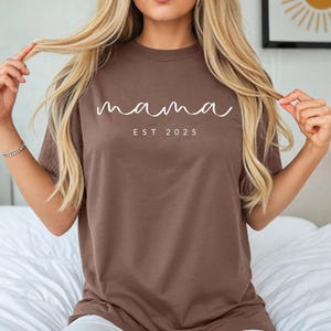 May include: A brown t-shirt with the text "mama est 2025" in white.