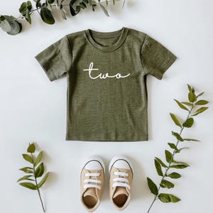 May include: Olive green toddler t-shirt with the word "two" in white script. The shirt is paired with tan canvas sneakers with white laces. Greenery surrounds the shirt and shoes on a white background.