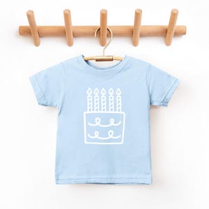 May include: A light blue t-shirt with a white outline of a birthday cake with candles on it.