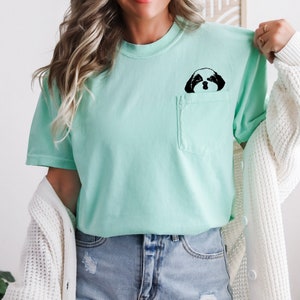 May include: A light green t-shirt with a black and white image of a dog's face peeking out of a pocket.
