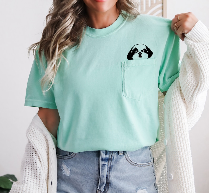 May include: A light green t-shirt with a black and white image of a dog's face peeking out of a pocket.
