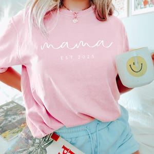 May include: Pink t-shirt with the text "mama est 2025" in white lettering.