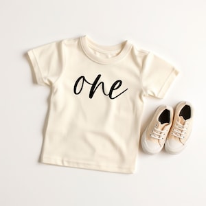 May include: A cream colored short-sleeved t-shirt with the word "one" written in black cursive lettering. The shirt is laying flat on a white surface next to a pair of cream colored sneakers.