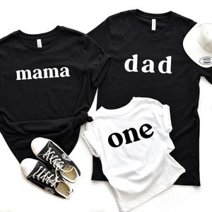 May include: Black t-shirts with the words "mama" and "dad" in white, along with a white t-shirt that says "one" in black. A white baseball cap and black sneakers are also in the image. Family matching apparel.