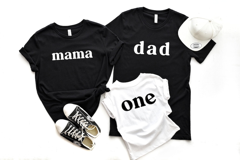 May include: Black t-shirts with the words "mama" and "dad" in white, along with a white t-shirt that says "one" in black. A white baseball cap and black sneakers are also in the image. Family matching apparel.