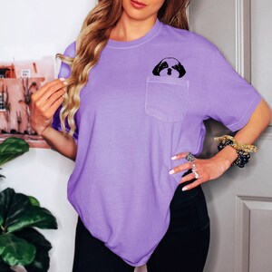 May include: A purple t-shirt with a small pocket on the left side. A black and white image of a dog's face is printed on the pocket.