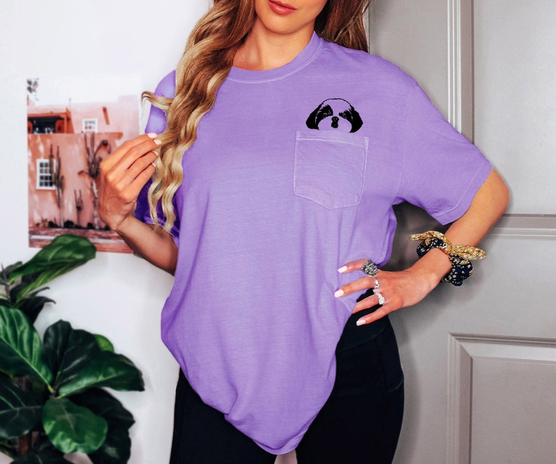 May include: A purple t-shirt with a small pocket on the left side. A black and white image of a dog's face is printed on the pocket.