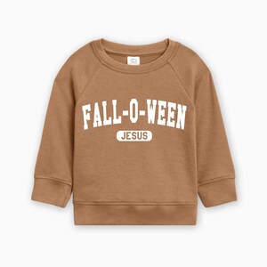 May include: A brown sweatshirt with the text "FALL-O-WEEN" in white block letters, with "JESUS" below. The long-sleeved top is designed for children.