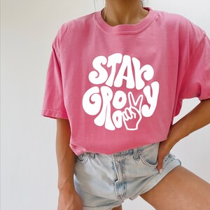 May include: A pink t-shirt with a white graphic that says "Stay Groovy" with a peace sign.