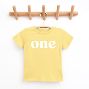 May include: A yellow t-shirt with the word "one" printed in white, hanging from a wooden peg rack. The shirt is a solid color and has short sleeves. The rack has five pegs.