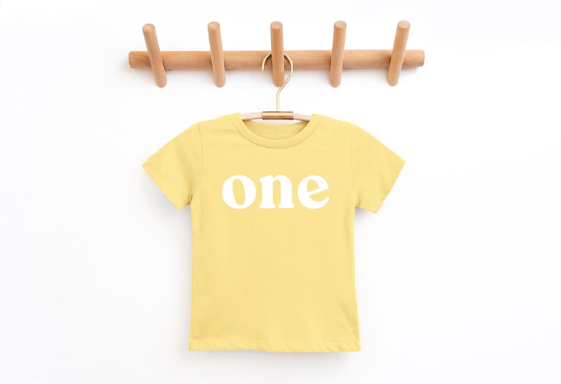May include: A yellow t-shirt with the word "one" printed in white, hanging from a wooden peg rack. The shirt is a solid color and has short sleeves. The rack has five pegs.