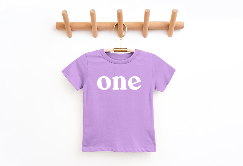 May include: A lilac-colored t-shirt with the word "one" printed in white, hanging on a wooden coat rack. The shirt is made of a soft material and is likely for a child's first birthday.