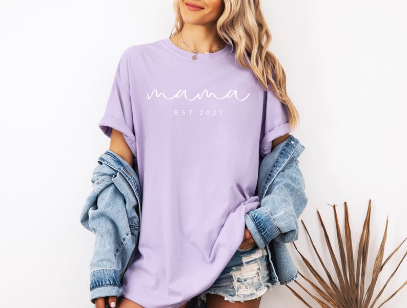 May include: A lavender t-shirt with the text "mama est 2023" in white.
