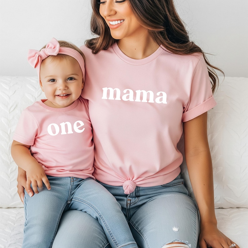 May include: Matching pink t-shirts with white text that reads "mama" and "one". The child wears a matching pink headband and light blue jeans. The adult is wearing light blue jeans.