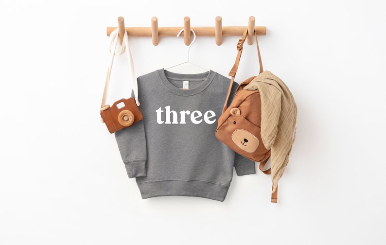 Puede incluir: Una sudadera gris con la palabra "three" impresa en blanco en la parte delantera. La sudadera est&aacute; colgada en un estante de madera con una mochila marr&oacute;n y una c&aacute;mara de juguete de madera.