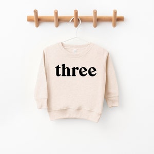 Puede incluir: Una sudadera de manga larga beige con la palabra "three" impresa en negro en la parte delantera.