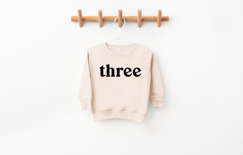 Puede incluir: Una sudadera de manga larga beige con la palabra "three" impresa en negro en la parte delantera.