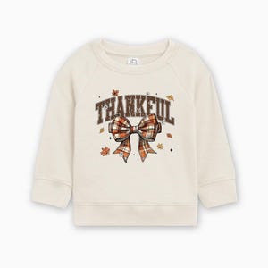 May include: Cream-colored sweatshirt with the word "THANKFUL" in brown block letters and a plaid bow graphic. The bow features shades of orange, brown, and white. Small leaf illustrations are scattered around the text and bow.