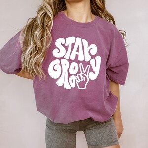May include: A purple t-shirt with white text that says "Stay Groovy" in a retro font with a peace sign. The shirt is being worn by a person with long blonde hair.