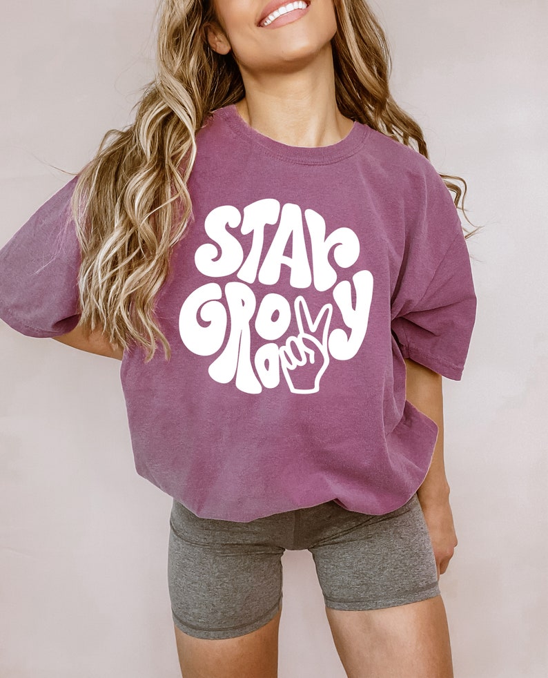 May include: A purple t-shirt with white text that says "Stay Groovy" in a retro font with a peace sign. The shirt is being worn by a person with long blonde hair.