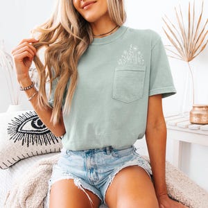 May include: A light green t-shirt with a pocket on the chest, featuring a white floral design. The shirt is paired with denim shorts. The overall aesthetic is casual and relaxed, suitable for everyday wear.