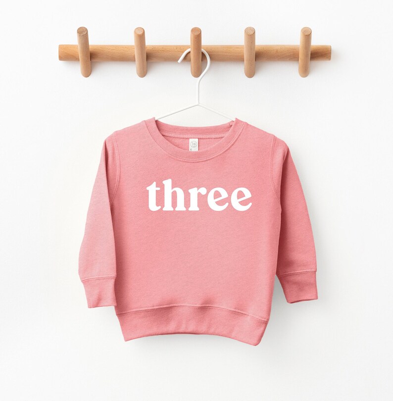Puede incluir: Una sudadera rosa de manga larga con la palabra "three" impresa en blanco en la parte delantera.