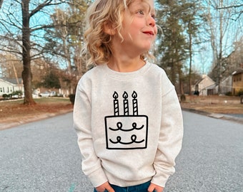 May include: A child wearing a light beige sweatshirt with a black and white birthday cake graphic. The cake has four candles with flames on top. The child is wearing blue jeans and has blonde hair.