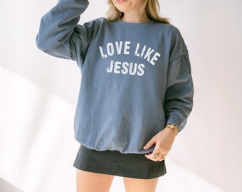 May include: A blue crew neck sweatshirt with the words "LOVE LIKE JESUS" printed in white. The sweatshirt is worn with a black skirt and a white baseball cap. The person is wearing sunglasses and gold jewellery.