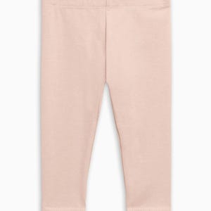 May include: A pair of solid light pink leggings featuring a high waistband and a fitted silhouette. The garment is constructed from a smooth, opaque fabric with visible stitching along the waist and the hem of each leg, providing a clean finish.