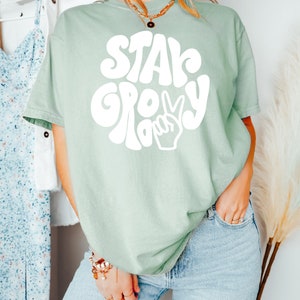 May include: A light green t-shirt with a white graphic that says "Stay Groovy" in a retro font with a peace sign.