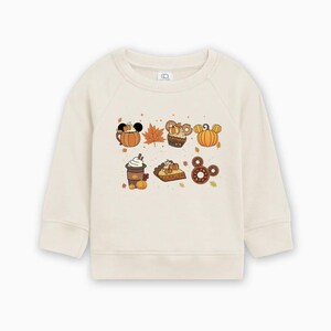 May include: Cream-colored sweatshirt featuring a colorful graphic of fall-themed food and leaf illustrations. The graphic includes a pumpkin spice latte, pie, donuts, and pumpkins with Mickey Mouse ears. A cozy, seasonal design.