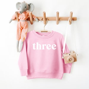 Puede incluir: Sudadera rosa para ni&ntilde;os con la palabra "three" impresa en blanco en la parte delantera.