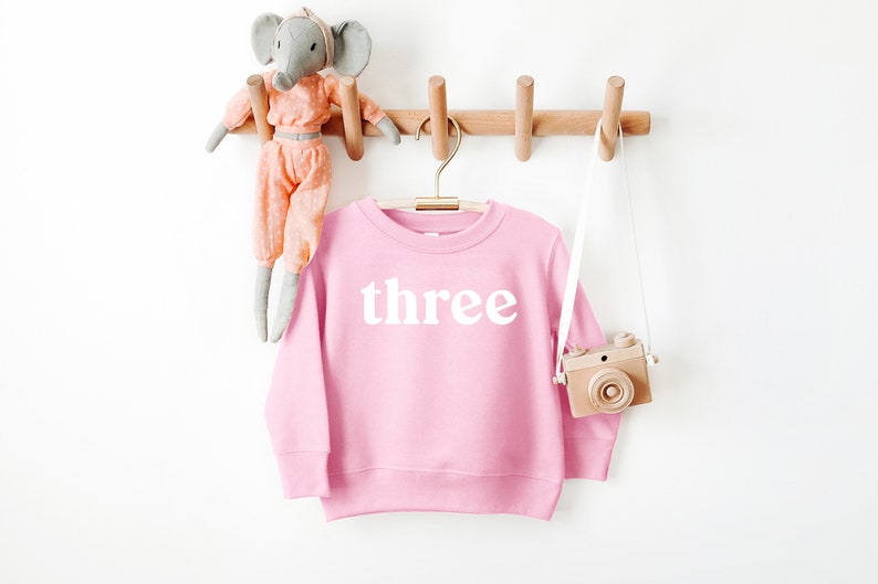Puede incluir: Sudadera rosa para ni&ntilde;os con la palabra "three" impresa en blanco en la parte delantera.