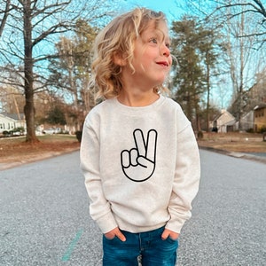 May include: A young child wearing a light beige sweatshirt with a black peace sign graphic. The child is standing on a paved road with trees in the background.