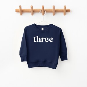 Puede incluir: Una sudadera de manga larga azul marino con la palabra "three" impresa en blanco en la parte delantera.