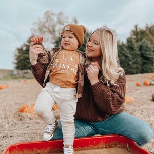 May include: A woman and a young child are standing in a pumpkin patch. The child is wearing a brown and white plaid shirt, a tan shirt with the text "Farm Fresh Pumpkins $5 Each", and white pants. The woman is wearing a brown sweater and blue jeans. They are standing in front of a red wagon.