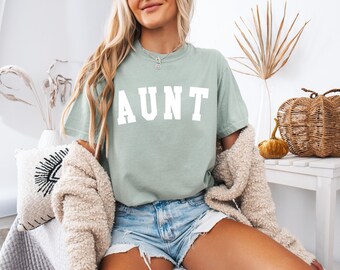 May include: A woman wearing a light green t-shirt with the word "AUNT" printed in white on the front. She is wearing denim shorts and a cream-colored cardigan.