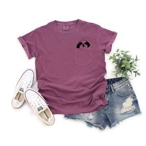 May include: A mauve t-shirt with a front pocket featuring a black and white illustration of a dog's face peeking out. The t-shirt is paired with denim shorts and white sneakers.