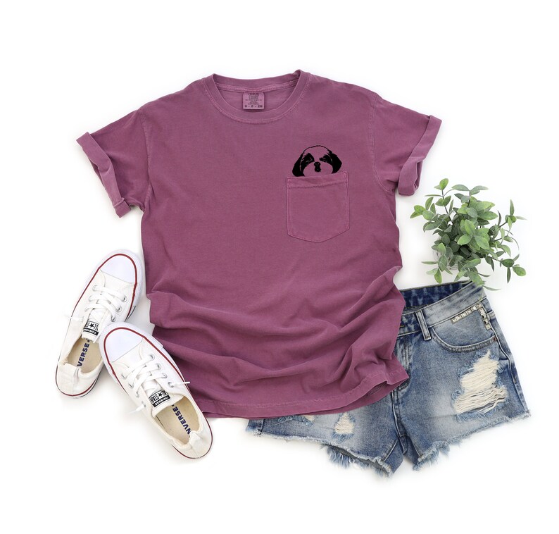 May include: A mauve t-shirt with a front pocket featuring a black and white illustration of a dog's face peeking out. The t-shirt is paired with denim shorts and white sneakers.