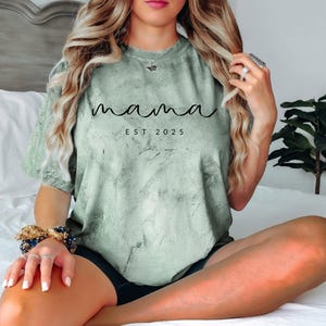 May include: A light green t-shirt with a distressed, marbled effect. The shirt has the text "mama est 2025" printed in black.