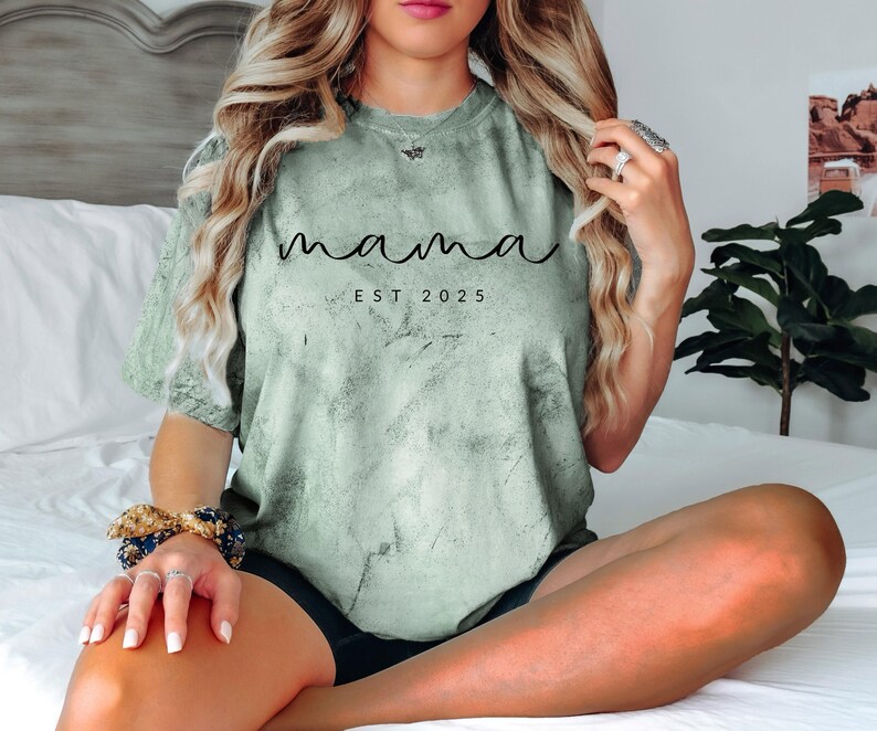 May include: A light green t-shirt with a distressed, marbled effect. The shirt has the text "mama est 2025" printed in black.