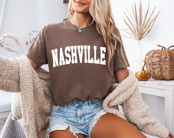 May include: A brown t-shirt with the word "NASHVILLE" printed in white on the front. The shirt is being worn by a person with long blonde hair and is paired with denim shorts and a cream-colored cardigan.
