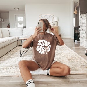 May include: A woman wearing a brown t-shirt with the text "Stay Groovy" in white. The text is in a retro font with a peace sign.