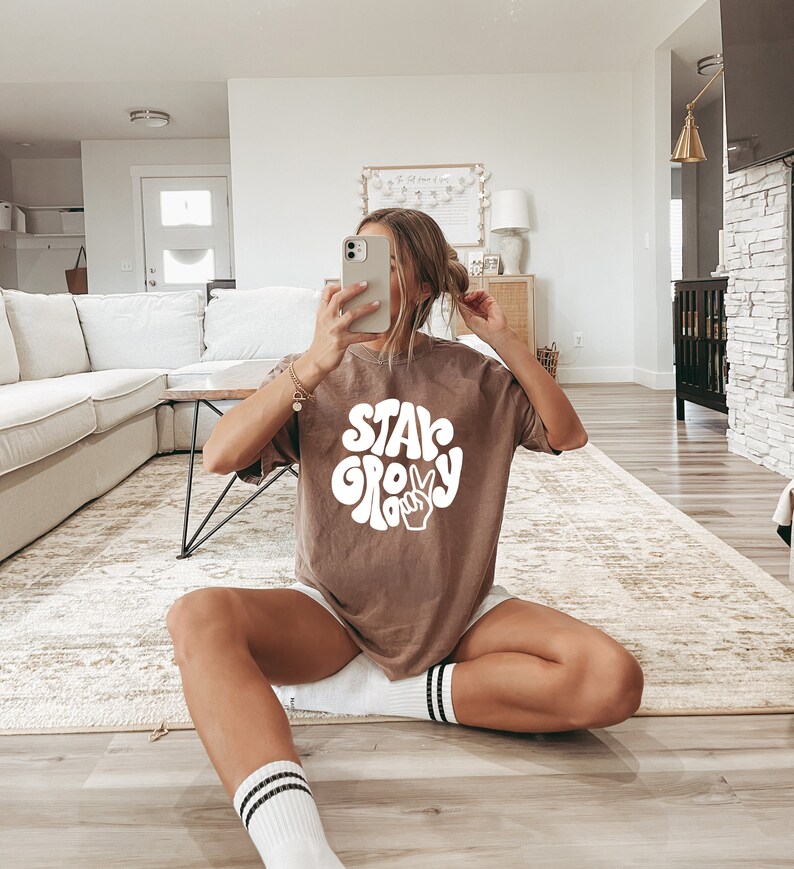 May include: A woman wearing a brown t-shirt with the text "Stay Groovy" in white. The text is in a retro font with a peace sign.