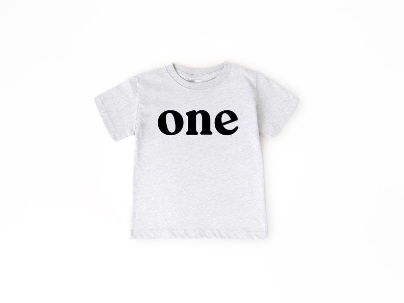 May include: A light gray toddler t-shirt with the word "one" printed in bold black letters across the front. The shirt has short sleeves and a crew neck.