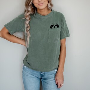 May include: A woman wearing a green t-shirt with a small black dog graphic on the pocket. She is wearing blue jeans and has long blonde hair.