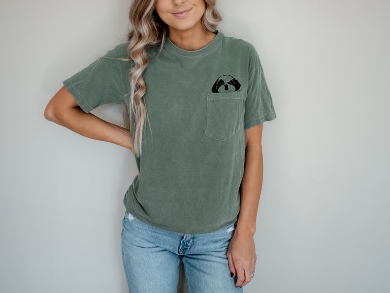 May include: A woman wearing a green t-shirt with a small black dog graphic on the pocket. She is wearing blue jeans and has long blonde hair.