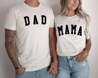 May include: A couple wearing white t-shirts with the words "DAD" and "MAMA" printed in black. They are holding hands and smiling.