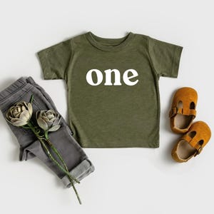 May include: Olive green toddler t-shirt with the word "one" in white block letters. The shirt is paired with gray pants, brown leather shoes, and decorative flowers. A stylish outfit for a first birthday celebration.